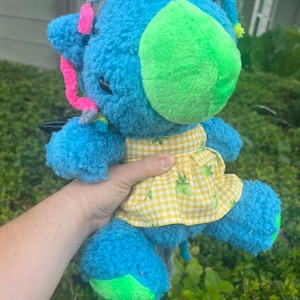 May include: A blue plush toy with a green nose and yellow and white checkered skirt. The toy has a long tail and is decorated with colorful yarn.