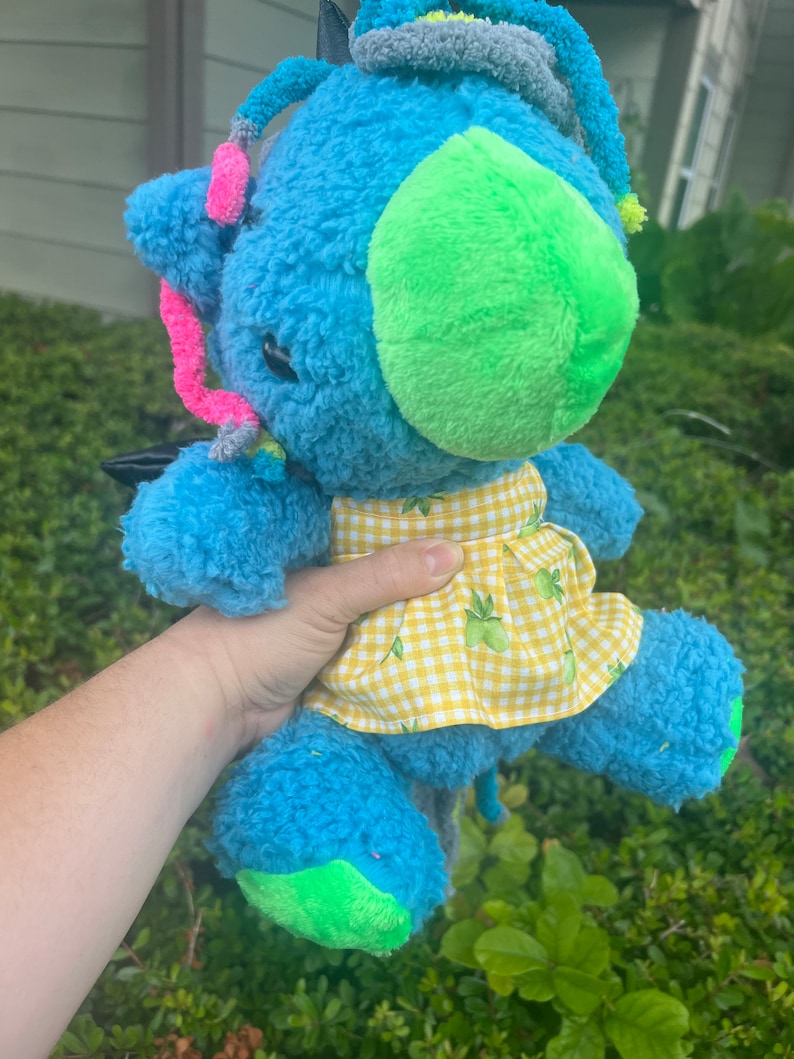 May include: A blue plush toy with a green nose and yellow and white checkered skirt. The toy has a long tail and is decorated with colorful yarn.