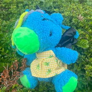 May include: A blue plush toy with green and yellow accents. The toy has a yellow and green gingham dress and black wings. The toy is sitting on a bed of green grass.
