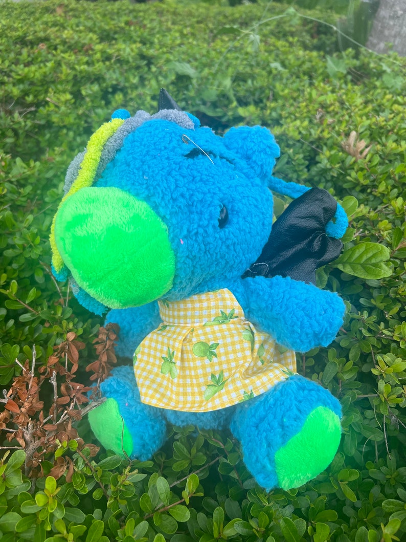 May include: A blue plush toy with green and yellow accents. The toy has a yellow and green gingham dress and black wings. The toy is sitting on a bed of green grass.