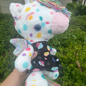 May include: White plush unicorn toy with colorful polka dots and a rainbow mane and tail. The unicorn is wearing a black dress with a cupcake print.