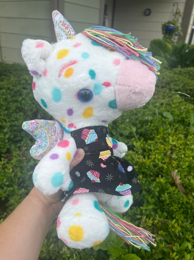 May include: White plush unicorn toy with colorful polka dots and a rainbow mane and tail. The unicorn is wearing a black dress with a cupcake print.
