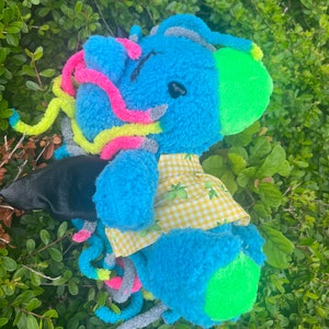 May include: A blue plush toy with a yellow and white checkered shirt. The toy has a green and gray tail and a green ear. The toy is sitting in a bed of green leaves.