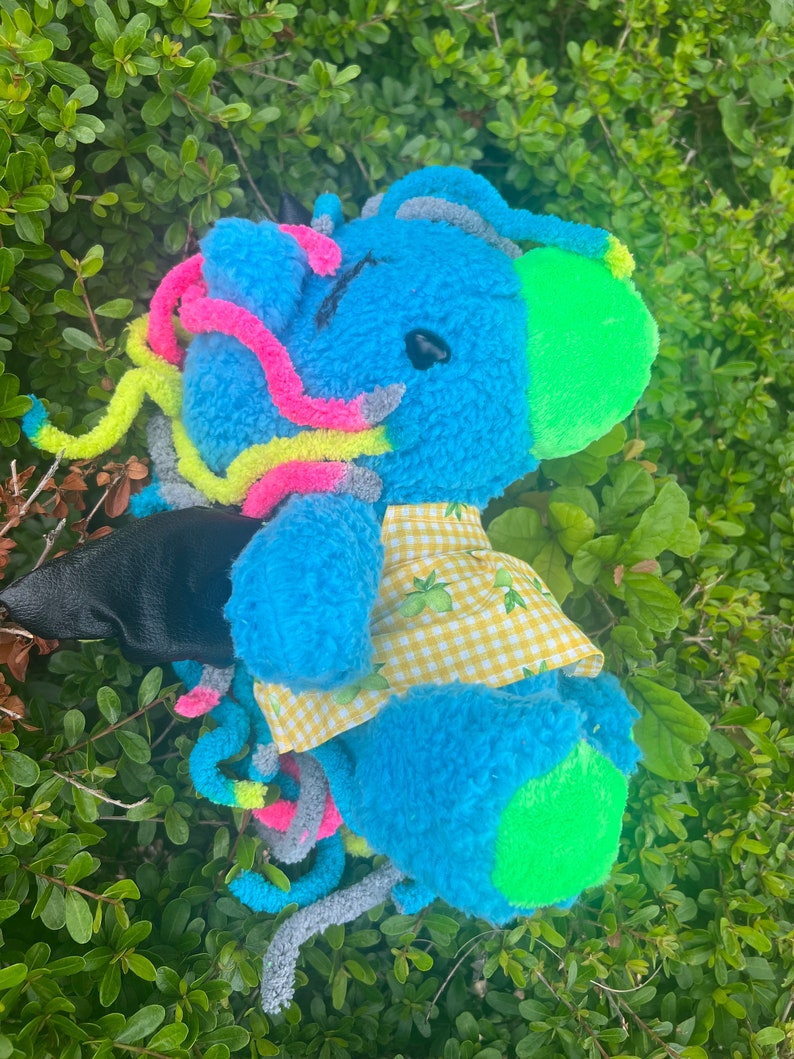 May include: A blue plush toy with a yellow and white checkered shirt. The toy has a green and gray tail and a green ear. The toy is sitting in a bed of green leaves.