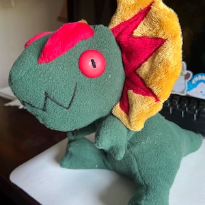 May include: Green plush dinosaur toy with a red and yellow crest, one large red button eye, and a stitched smile.