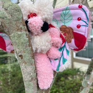 Op de afbeelding: Een roze en wit pluche speelgoed met vlinder vleugels. De vleugels zijn roze met een bloemenpatroon en rode bessen. Het speelgoed zit op een tak.
