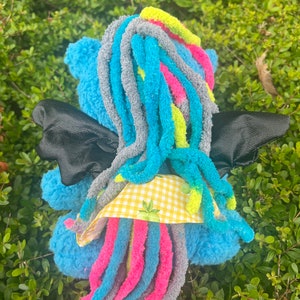 May include: A blue plush toy with long, colorful yarn hair, black wings, and a yellow and white checkered fabric detail. The toy is sitting on a bed of green foliage.