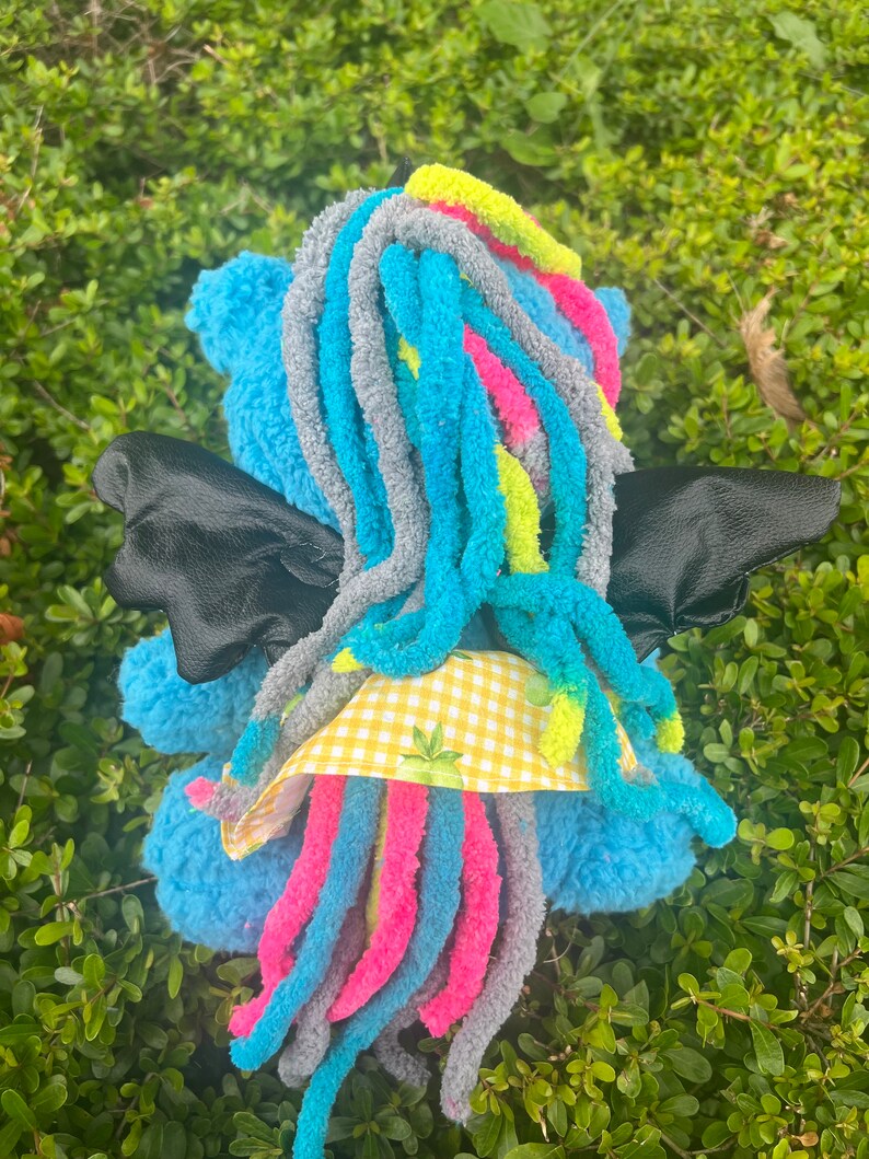 May include: A blue plush toy with long, colorful yarn hair, black wings, and a yellow and white checkered fabric detail. The toy is sitting on a bed of green foliage.