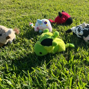 May include: Five plush toys in the shape of farm animals. A brown and white cow, a green and black cow, a white and pink unicorn, a red bull, and a brown sheep.