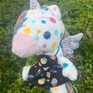 May include: A white plush unicorn toy with a rainbow mane and tail. The unicorn is wearing a black dress with a cupcake pattern. The unicorn has a blue eye and is covered in colorful polka dots.