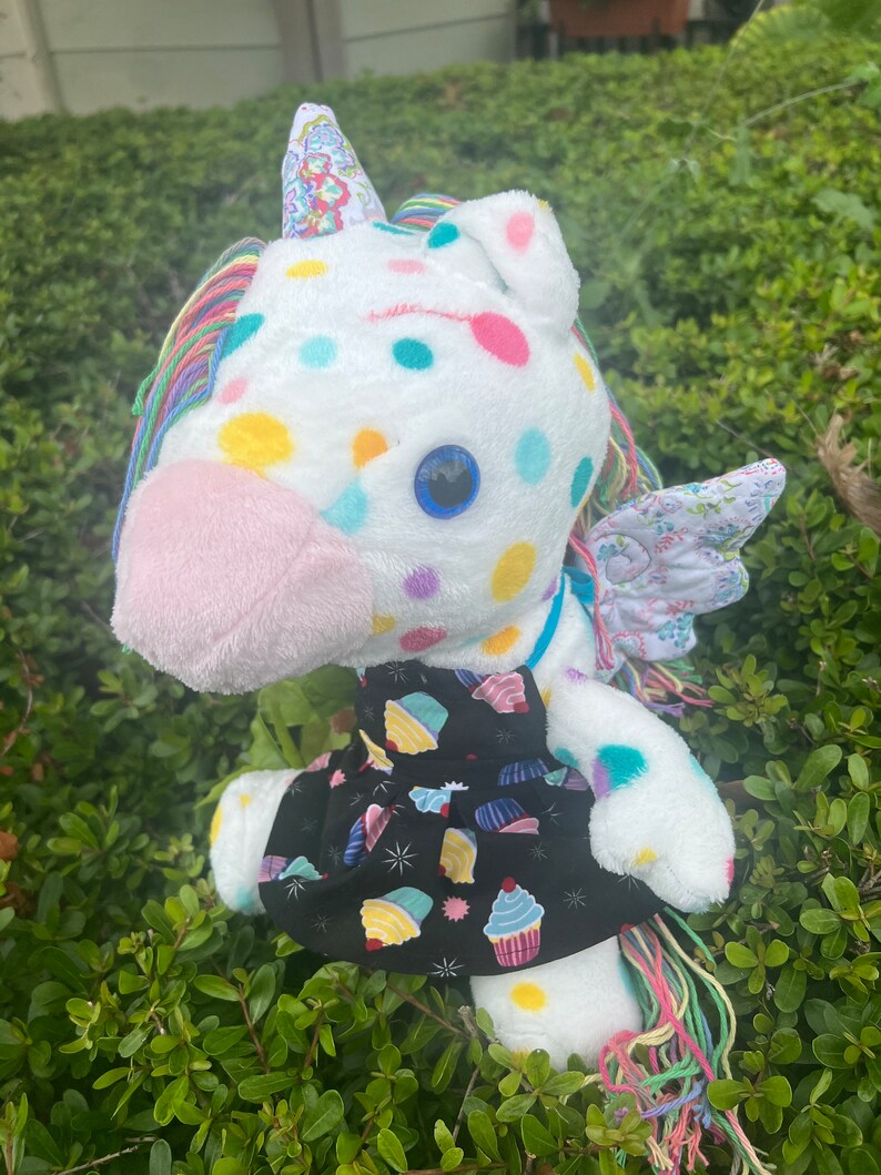 May include: A white plush unicorn toy with a rainbow mane and tail. The unicorn is wearing a black dress with a cupcake pattern. The unicorn has a blue eye and is covered in colorful polka dots.