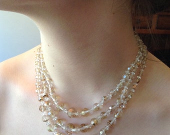 May include: A multi-strand necklace made of clear and gold-toned glass beads. The necklace is layered and has a delicate, feminine look.