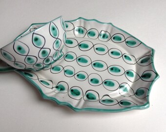 May include: A white ceramic teacup and saucer set. The set features a teal and black oval pattern with a scalloped edge. The teacup has a handle and sits on a matching oval-shaped saucer.