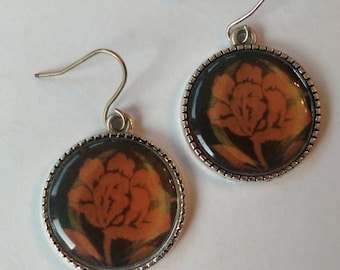 May include: A pair of silver earrings with a round, orange flower design. The earrings have a silver hook closure.