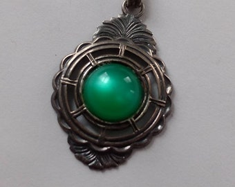 May include: A silver pendant necklace with a green gemstone set in a circular frame. The frame has a decorative design with intricate details.