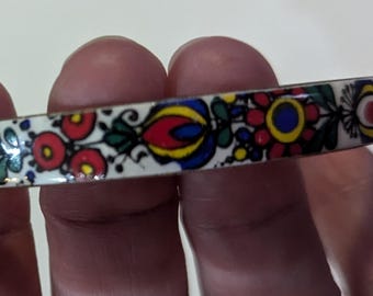 May include: A white enamel bangle bracelet with a colorful floral pattern. The design features red, yellow, blue, and green flowers and leaves. The bracelet is held in a hand.