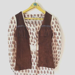 May include: A vintage outfit comprising a brown suede vest with fringing, worn over a cream blouse. The blouse has a button-down front and a paisley pattern in red and brown. The outfit is displayed on a yellow hanger.