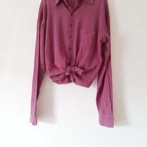 May include: A long-sleeved button-down shirt in a deep mauve color with a textured, crinkled fabric finish. The shirt features a classic collar, a single chest pocket, and is tied at the front waist. A wooden hanger is visible at the top.