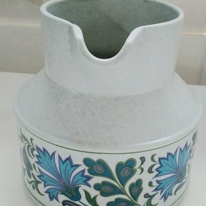 Op de afbeelding: Een witte keramische kan met een blauw en groen bloemenpatroon. De kan heeft een brede opening en een handvat.