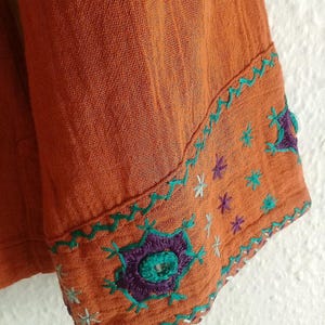 以下が含まれることがあります： オレンジ色の生地のスリーブのクローズアップ。刺繍のディテールが施されています。スリーブには、ティール、紫、白の花柄とスターバースト模様があります。生地は質感のある外観をしています。