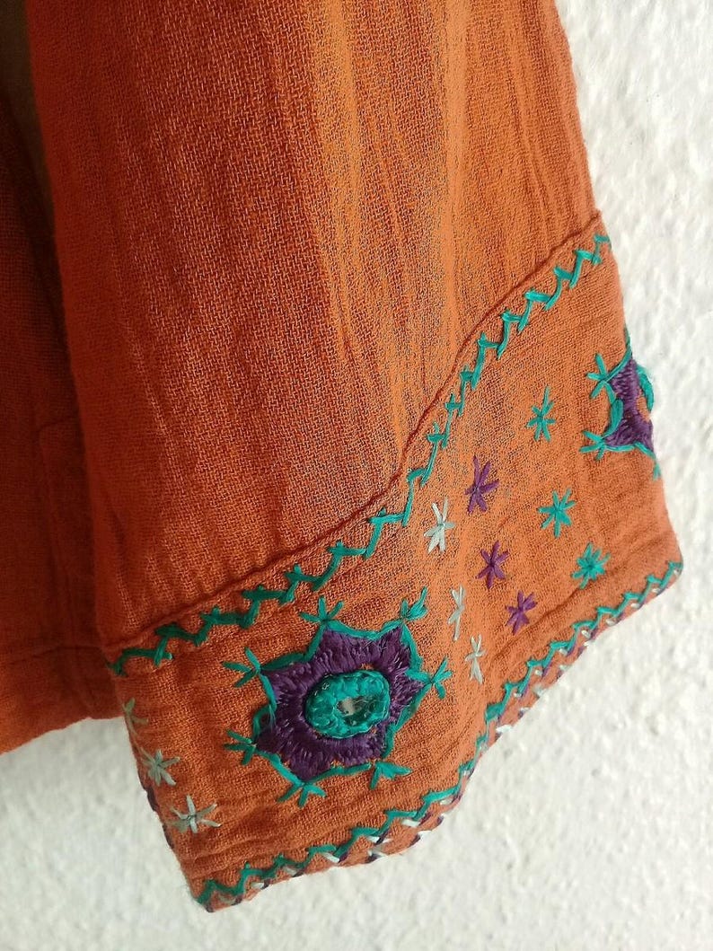 以下が含まれることがあります： オレンジ色の生地のスリーブのクローズアップ。刺繍のディテールが施されています。スリーブには、ティール、紫、白の花柄とスターバースト模様があります。生地は質感のある外観をしています。