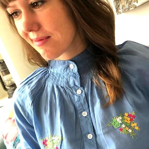 May include: A pale blue blouse with a high collar and white buttons. The blouse has embroidered floral designs in pink, yellow, green, and white. The top has a gathered neckline and long sleeves.