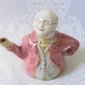 Può includere: Una teiera in ceramica a forma di persona che indossa una giacca rosa, una camicia bianca e dettagli dorati. La teiera ha un beccuccio a forma di braccio e un coperchio sulla testa.