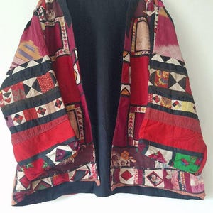 Può includere: Una giacca a maniche lunghe caratterizzata da un design patchwork con vari quadrati di tessuto rossi, neri e multicolori. Il capo presenta una fodera interna nera a tinta unita, un'apertura frontale ed è esposto su una gruccia in legno.