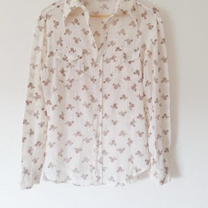 May include: A long-sleeved button-down shirt in an off-white color featuring a repeating brown floral pattern. The shirt includes a pointed collar, a front button placket, and two chest pockets with buttoned flaps, all displayed on a wooden hanger.