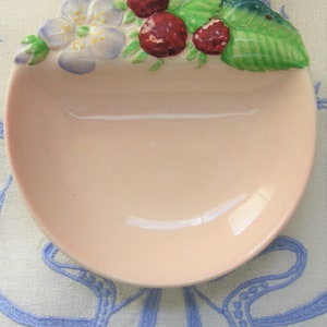 May include: A pink ceramic dish with a raised design of strawberries and a flower. The dish is decorated with green leaves and has a scalloped edge.
