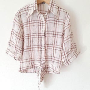 May include: A button-down shirt with a white and brown plaid pattern, featuring a pointed collar, three-quarter length sleeves with folded cuffs, and a tie-front hem detail. The garment is made of a textured fabric and hangs on a wooden hanger.