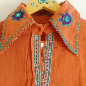 以下が含まれることがあります： オレンジ色のボタンダウンシャツ、刺繍入り。 襟には紫とターコイズの花柄刺繍が施されています。 ターコイズと白のステッチがフロントを開いています。 シャツは、青い車のデザインの木製ハンガーにかかっています。
