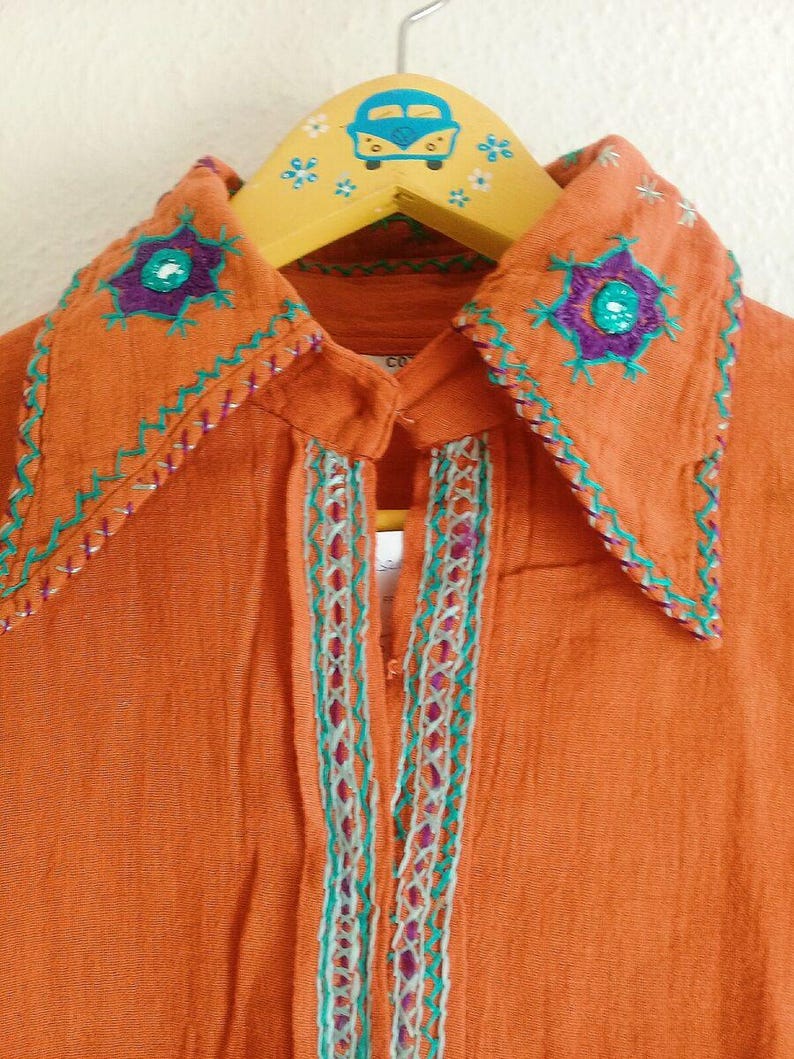 以下が含まれることがあります： オレンジ色のボタンダウンシャツ、刺繍入り。 襟には紫とターコイズの花柄刺繍が施されています。 ターコイズと白のステッチがフロントを開いています。 シャツは、青い車のデザインの木製ハンガーにかかっています。