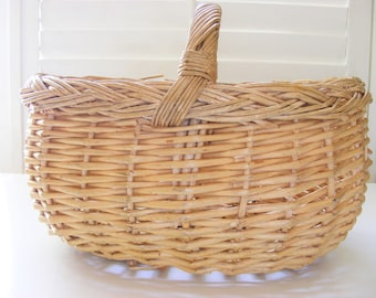 May include: A large, oval-shaped wicker basket with a woven handle. The basket is made of natural, light brown wicker.