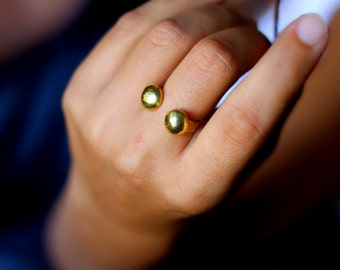 Puede incluir: Primer plano de una mano con dos anillos de oro. Cada anillo presenta una piedra redondeada de color dorado. Los anillos son sencillos y elegantes, con un acabado pulido. El fondo está borroso, centrando la atención en las joyas.