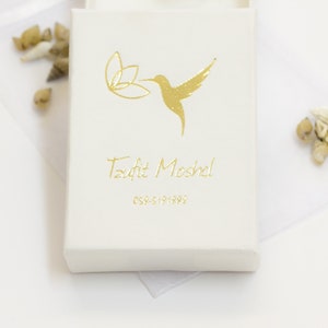 Puede incluir: Una caja blanca con un colibr&iacute; dorado y un dise&ntilde;o floral. El texto "Tzofit Moshel" y "054-5191892" est&aacute; impreso en dorado en la caja.