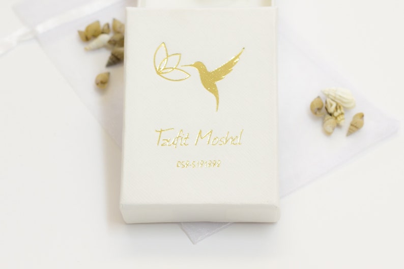 Puede incluir: Una caja blanca con un colibr&iacute; dorado y un dise&ntilde;o floral. El texto "Tzofit Moshel" y "054-5191892" est&aacute; impreso en dorado en la caja.