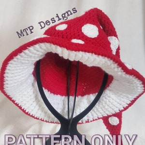 Könnte beinhalten: Ein roter und weißer gehäkelter Pilz-Hut mit weißen Punkten. Der Hut steht auf einem schwarzen Ständer. Der Text "MTP Designs" und "PATTERN ONLY" ist auf dem Bild zu sehen.