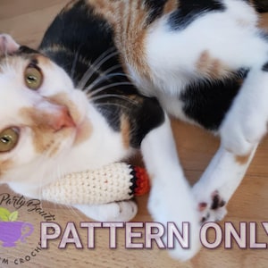 Puede incluir: Un gato calico con un juguete de gato de crochet blanco con detalles rojos y naranjas. El juguete de gato tiene forma de dedo. El texto "PATTERN ONLY" está superpuesto en la imagen.