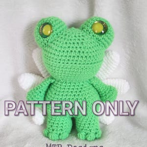 May include: A crocheted green frog with large yellow eyes and white wings. The frog has the text "PATTERN ONLY" and "MTP Designs" printed on the image.