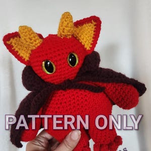 May include: A handmade red crochet dragon with yellow accents on the ears and horns, and large, glittery eyes. The dragon has a dark brown cape and the text "PATTERN ONLY" is visible. The dragon is being held by a person.