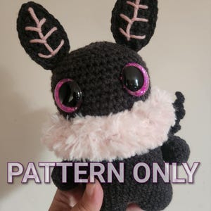 May include: Crochet pattern for a black amigurumi creature with pink ears and a pink fur collar. The creature has large black eyes with pink glitter. The text "PATTERN ONLY" is visible in the image.