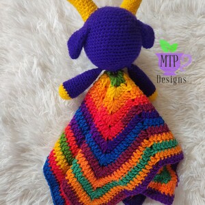 May include: A purple crocheted goat with yellow horns holding a rainbow crocheted blanket. The blanket has a chevron pattern and is made with different colors of yarn. The goat and blanket are on a white fluffy surface. MTP Designs is written on a green leaf.