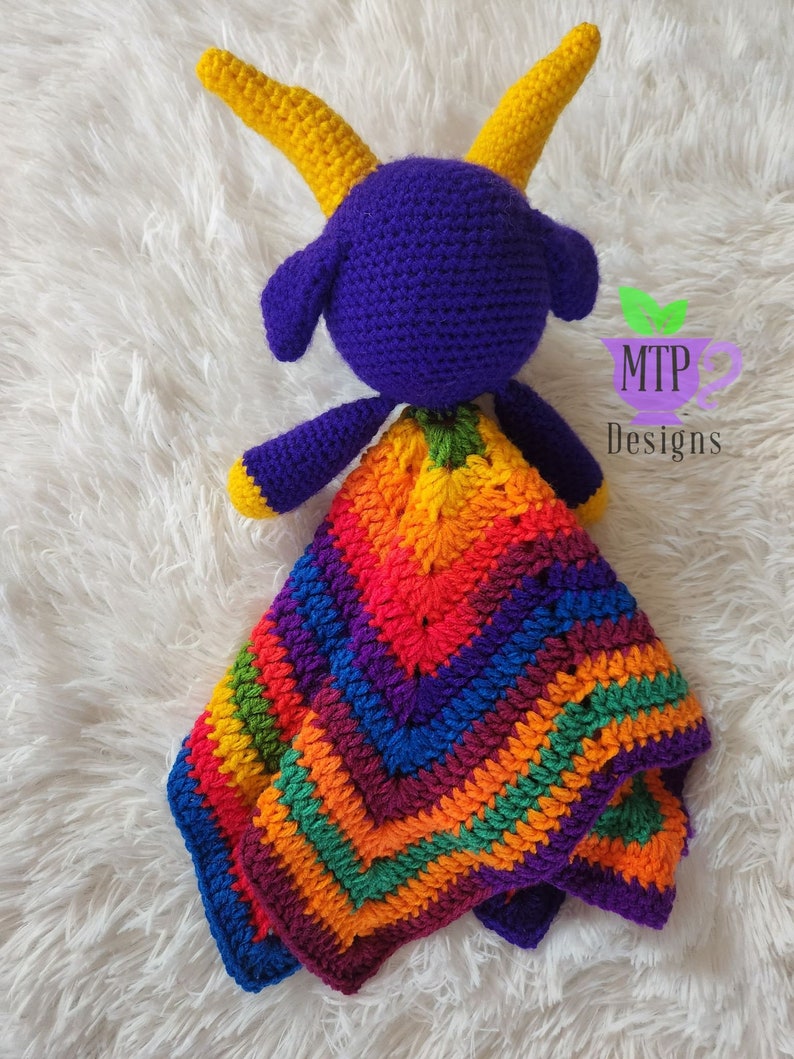 May include: A purple crocheted goat with yellow horns holding a rainbow crocheted blanket. The blanket has a chevron pattern and is made with different colors of yarn. The goat and blanket are on a white fluffy surface. MTP Designs is written on a green leaf.