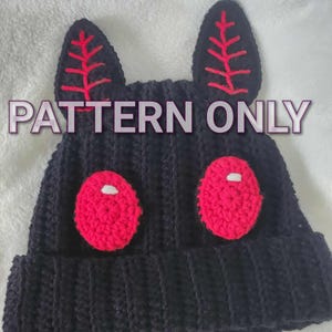 Op de afbeelding: Zwarte gehaakte muts met rode accenten. Voorzien van twee rode, ovaalvormige ogen en twee rechtopstaande oren met rode details. De tekst "PATTERN ONLY" en "MTP Designs" zijn zichtbaar.
