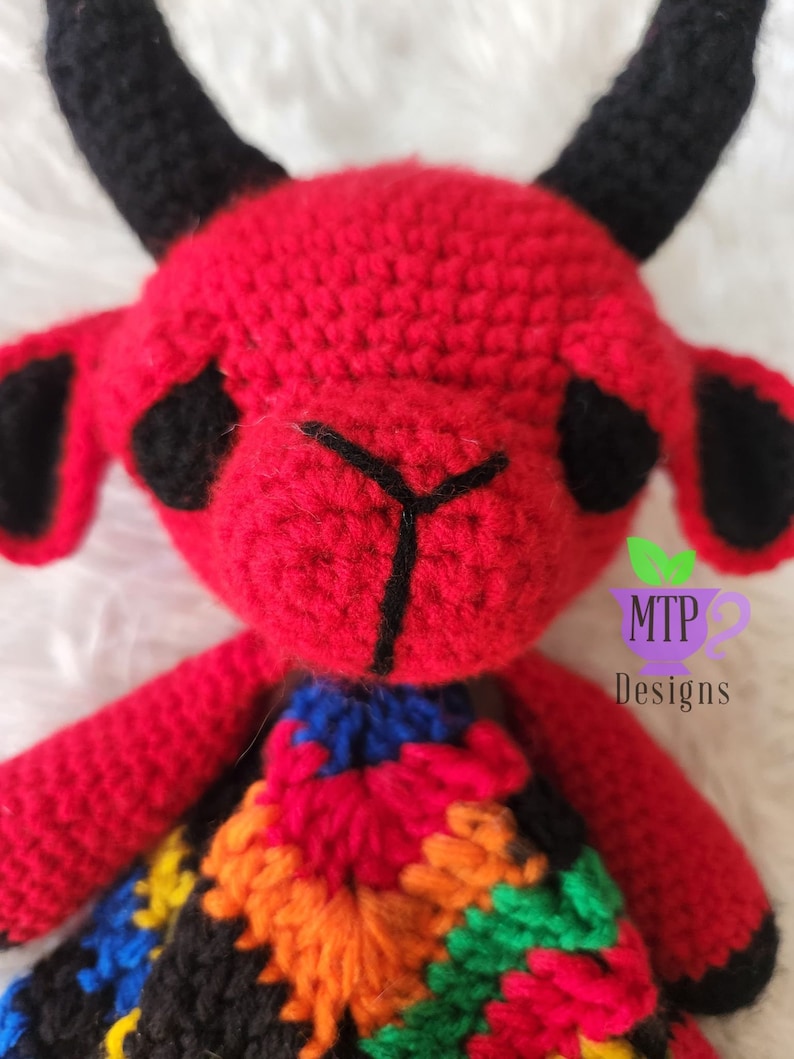 May include: A red crocheted devil with black horns and a colorful crocheted skirt. The devil has black eyes and a black stitched nose. The skirt is made of different colored squares. The image includes the text "MTP Designs".