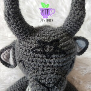 May include: A gray crocheted stuffed animal with horns and a black star on its forehead. The animal is wearing a multi-colored scarf.