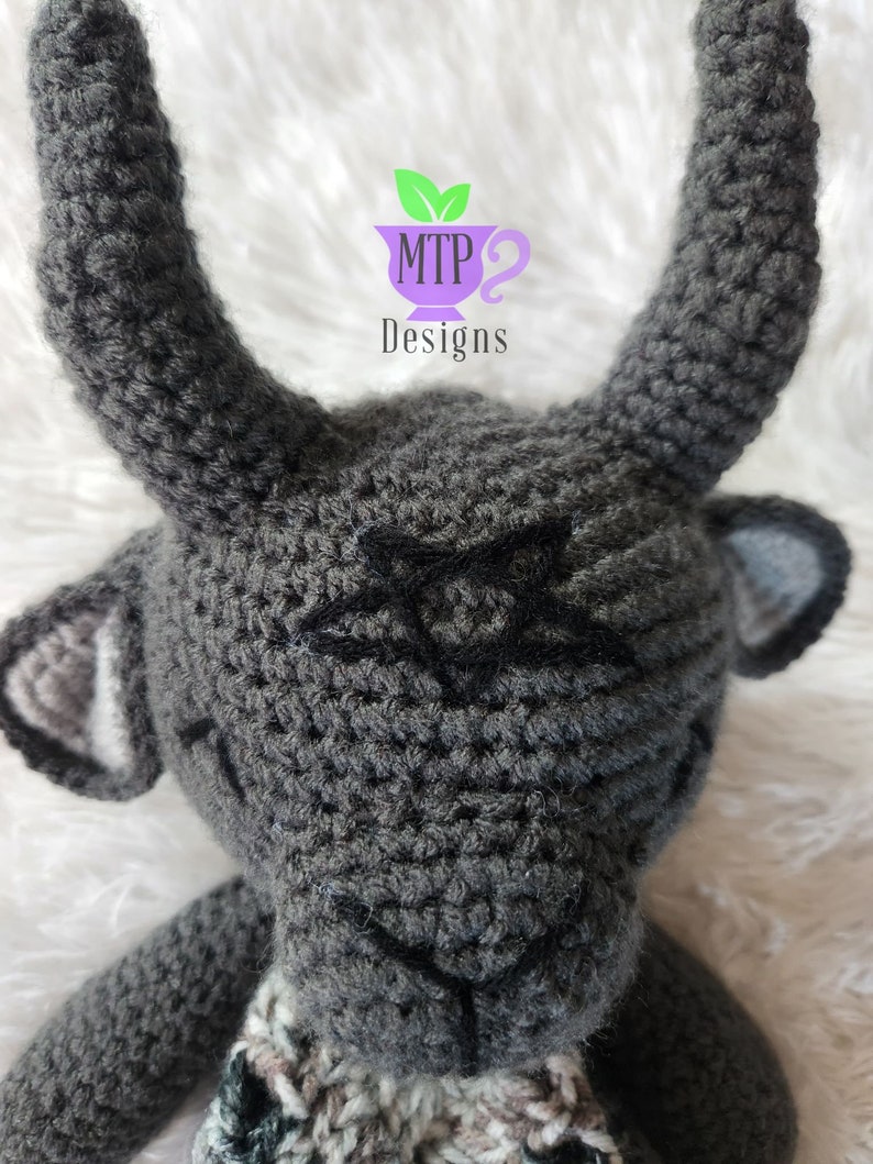 May include: A gray crocheted stuffed animal with horns and a black star on its forehead. The animal is wearing a multi-colored scarf.