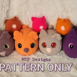 May include: A collection of seven crocheted stuffed animals in various colors, including purple, orange, gray, and pink. The animals have large eyes and are arranged in a row on a white fluffy surface. The text "MTP Designs PATTERN ONLY" is visible at the bottom of the image.