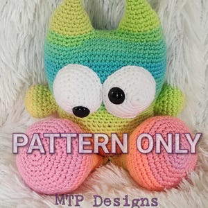 Puede incluir: Un juguete de monstruo de ganchillo hecho a mano en tonos verdes, azules y amarillos, con ojos blancos grandes y pupilas negras. El juguete tiene pies rosas y naranjas. El texto "PATTERN ONLY" y "MTP Designs" son visibles.